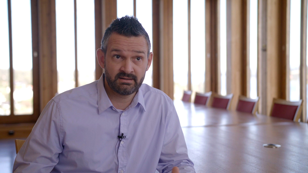Mann med mørkt hår, kort skjegg og blå skjorte sitter i et tomt møterom og snakker mot kamera.